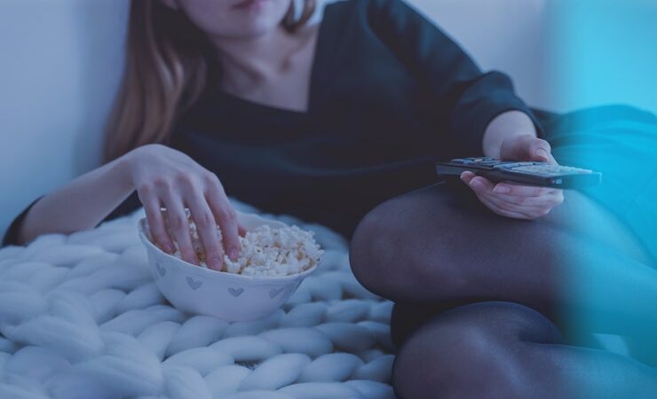 Woman laying on soft blanket watching tv holding a remote and eating popcorn