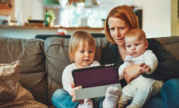 Mom holding her two children while looking at their tablet while sitting on their couch