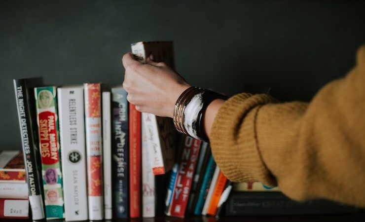 Woman picking a book out of shelf full used books while wearing brown sweater and bangles
