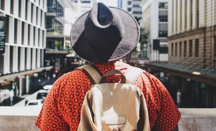 Young man wearing a black hat a back pack and an orange shirt looking out at the city view