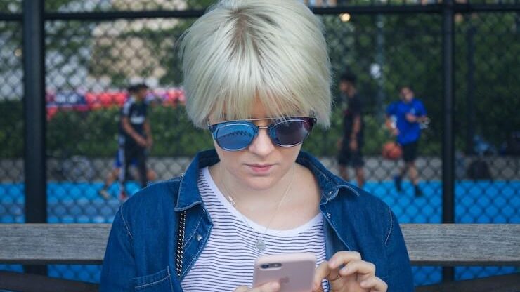 Blond girl sitting in a park wearing sunglasses using her cell phone