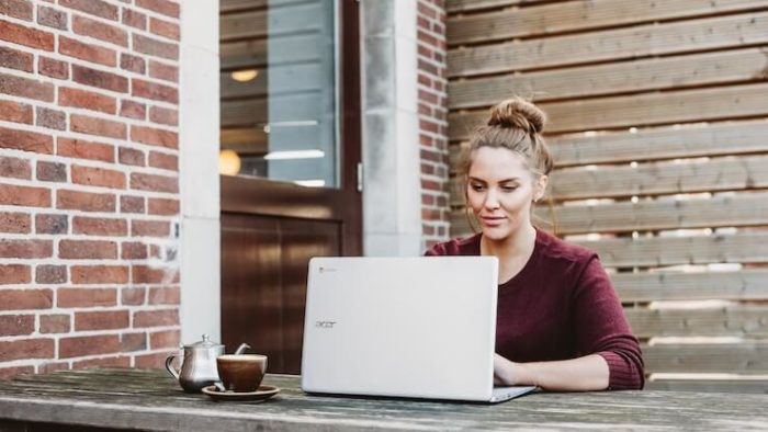 Woman with a messy bun working on an acer laptop outside on picnic table next to a brick wall