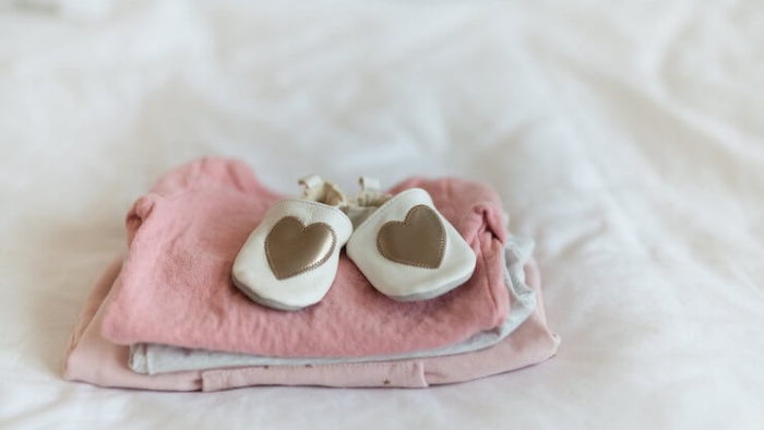Baby shoes with gold heart on them sitting on top of pink clothing folded on a bed