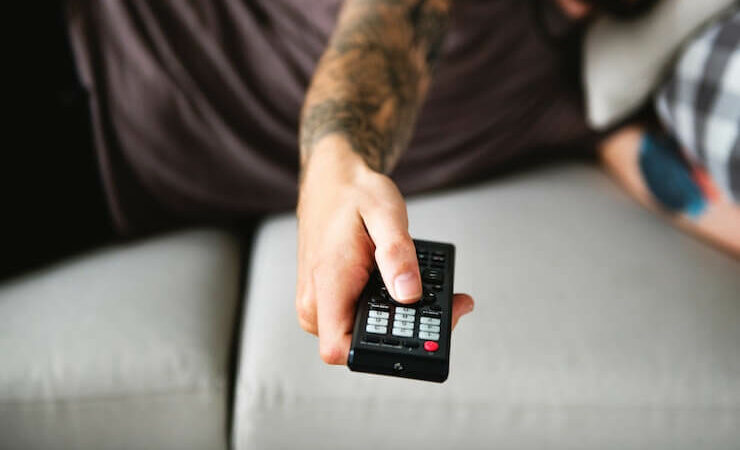 Man laying down on a grey couch changing the tv channel with a remote