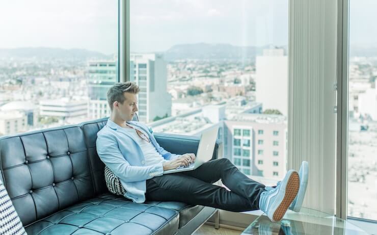 Man sitting on a couch using his laptop with an amazing view from the windows behind him