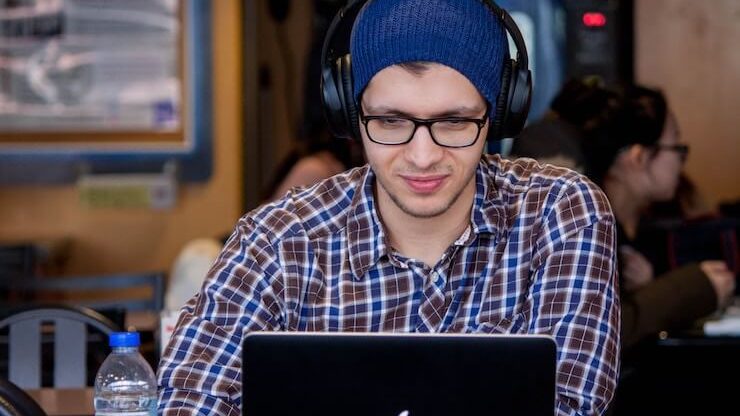 young man student wearing blue beanie using his computer wearing headphones in coffee shop